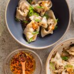 Hero Shot Wontons with Spicy Chili Sauce. 5 wontons in a black bowl, and 5 in a cream bowl, next to a small dish of diagonallly sliced green onione, and next to a small clear glass bowl with Spicy Chili OIl Sauce with a tiny wooden spoon.