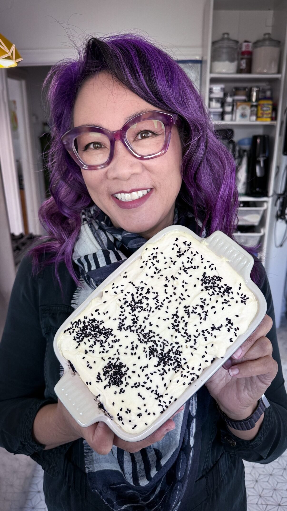 Carole holding a tray of black sesame tiramisu, up to the camera, so that you can see the cream topping sprinkled with black sesame seeds. Carole has large oversized purple glasses and curled shoulder length purple glasses hair.