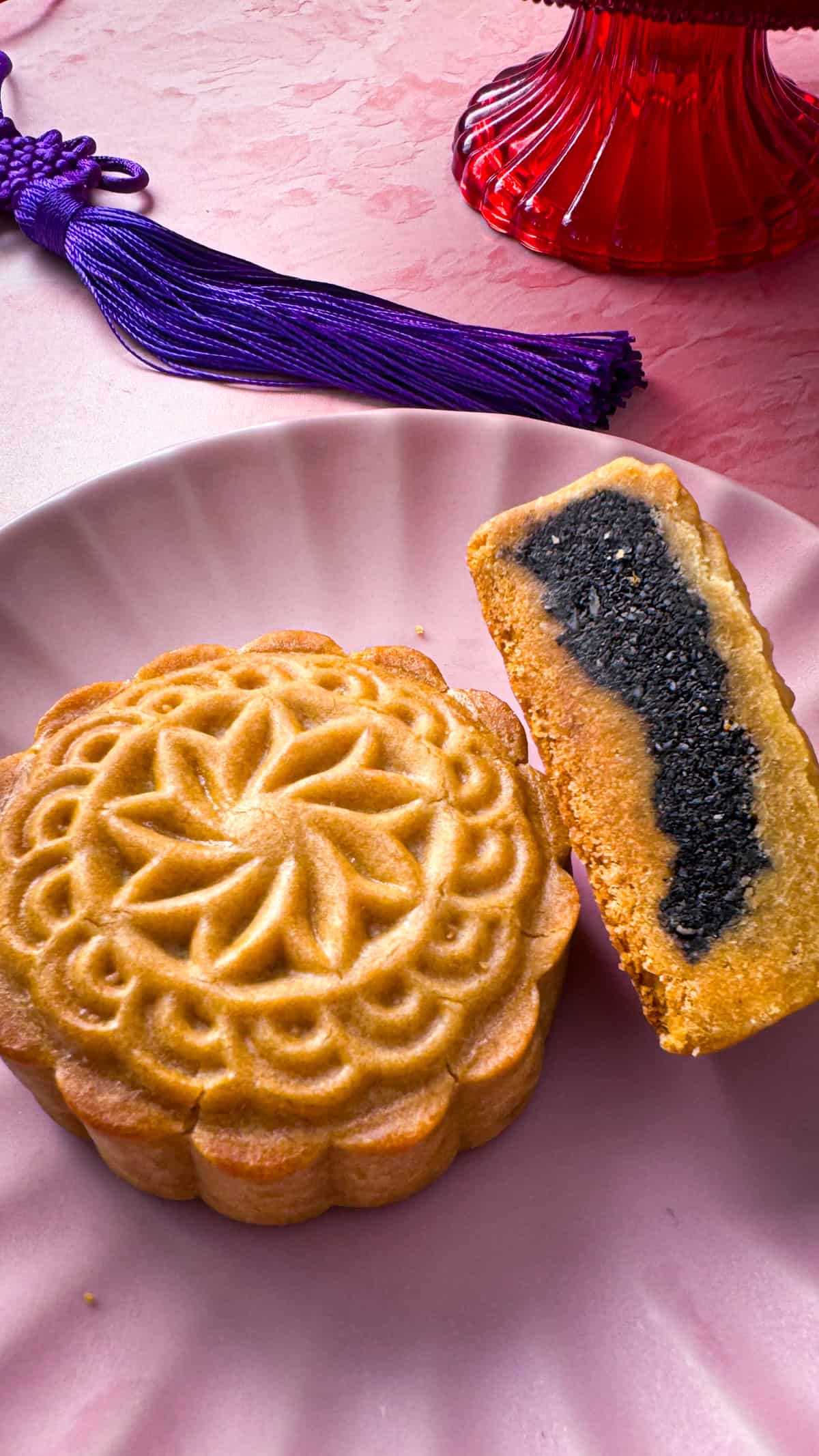 A cut view of half a Black Sesame Mooncake on a pink Plate, next to a full mooncake, showing the beautiful star print on the top of the cake.