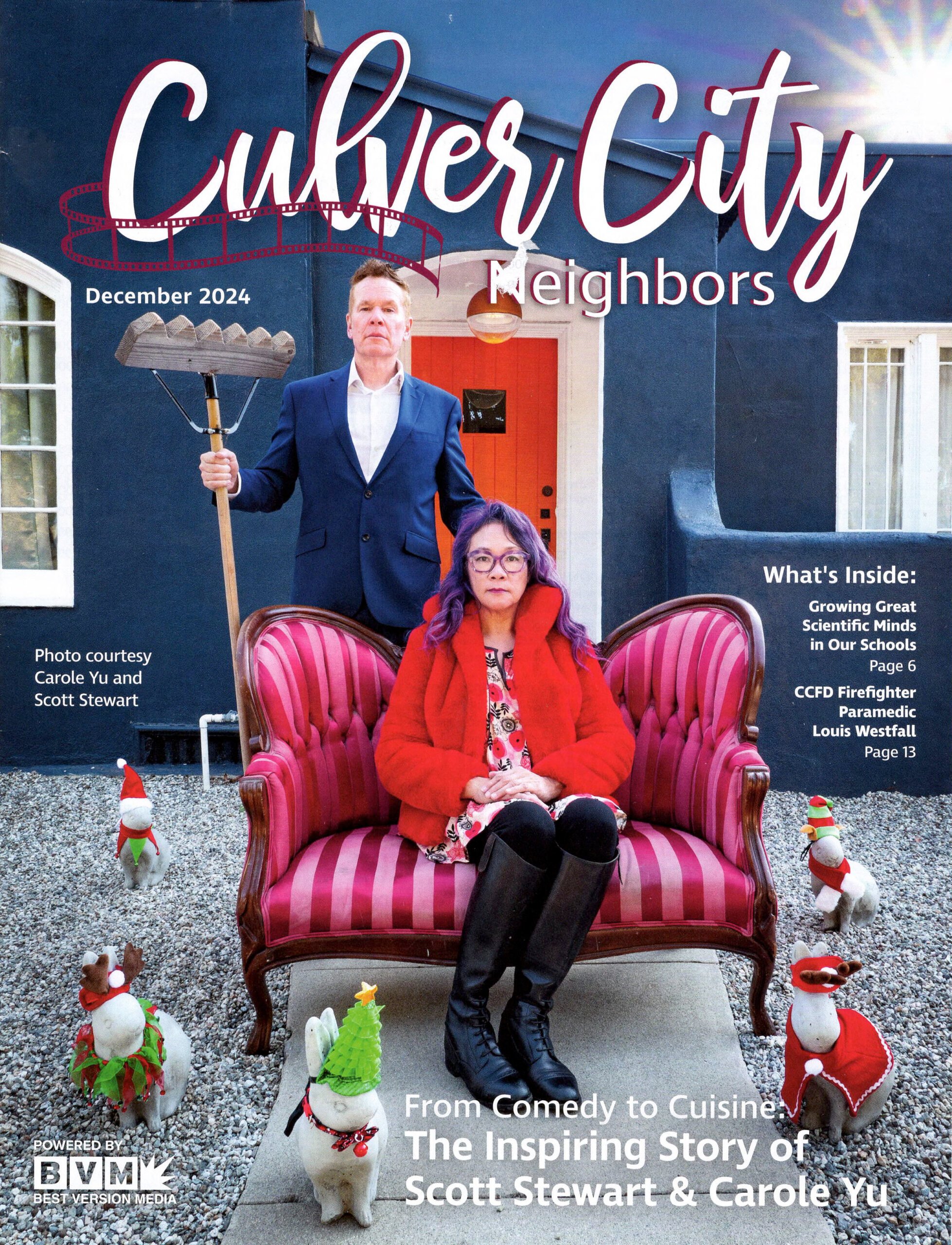 Cover of Dec 2024 Culver City Neighbors magazine with Scott in a blue suit holding a rake, and Carole in a red fur jacket, and black horse riding boots, sitting on a vintage red, and pink striped loveseat. They are in front of their home which is a dark Blue home, with white window frames, and a bright orange fron door.