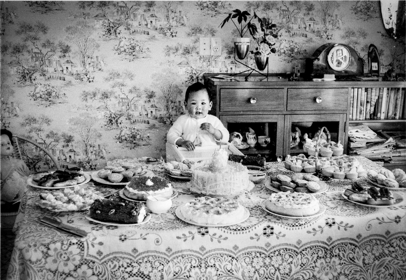 Black and White photo of Baby Carole at her first birthday party, sitting in front of a lace covered table, surrounded by 15 desserts, cake, pavlovas, chocolate log, tarts, butterfly cakes, madeleines, chocolate eclairs, and more.
