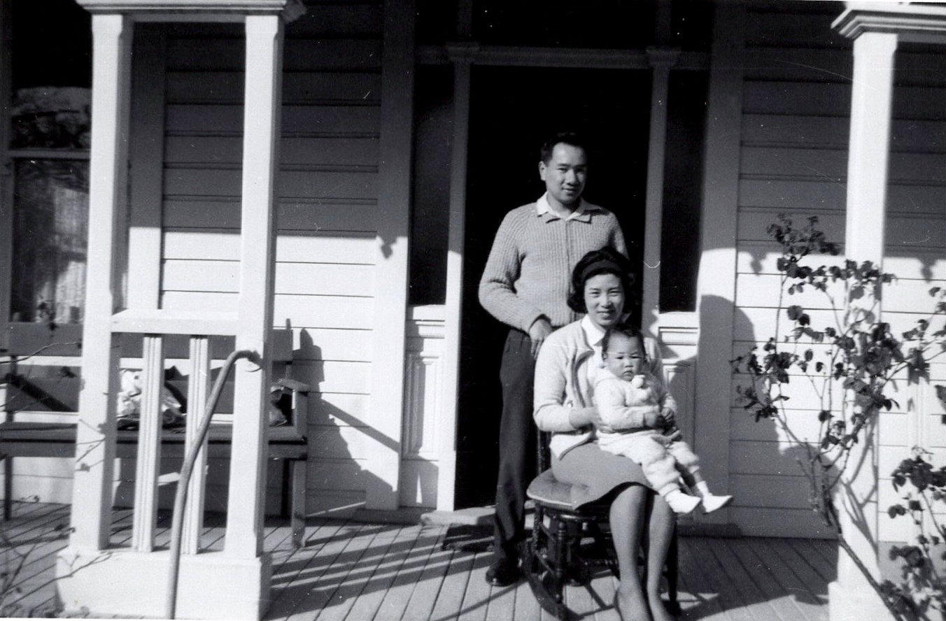 Baby Carole dressed in a hand made knitted pants, and sweater with pompoms, sitting on her mother, Nellie's lap, and father, Warren standing behind the chair. They are on the porch of an old house in Dunedin, New Zealand where Carole was born.