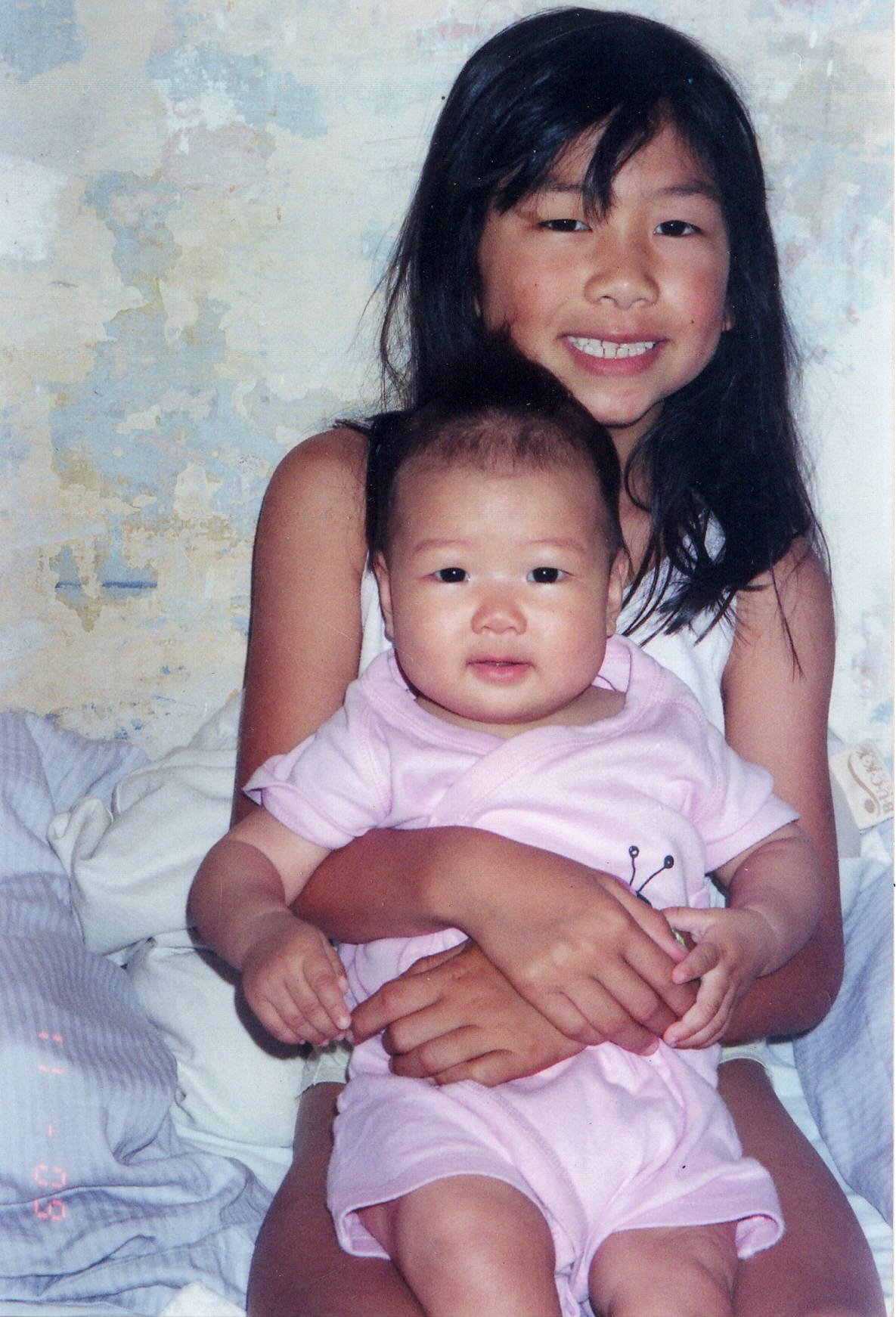 2001 Elena and Dara sitting on bed, with pastel mottled wall background.
