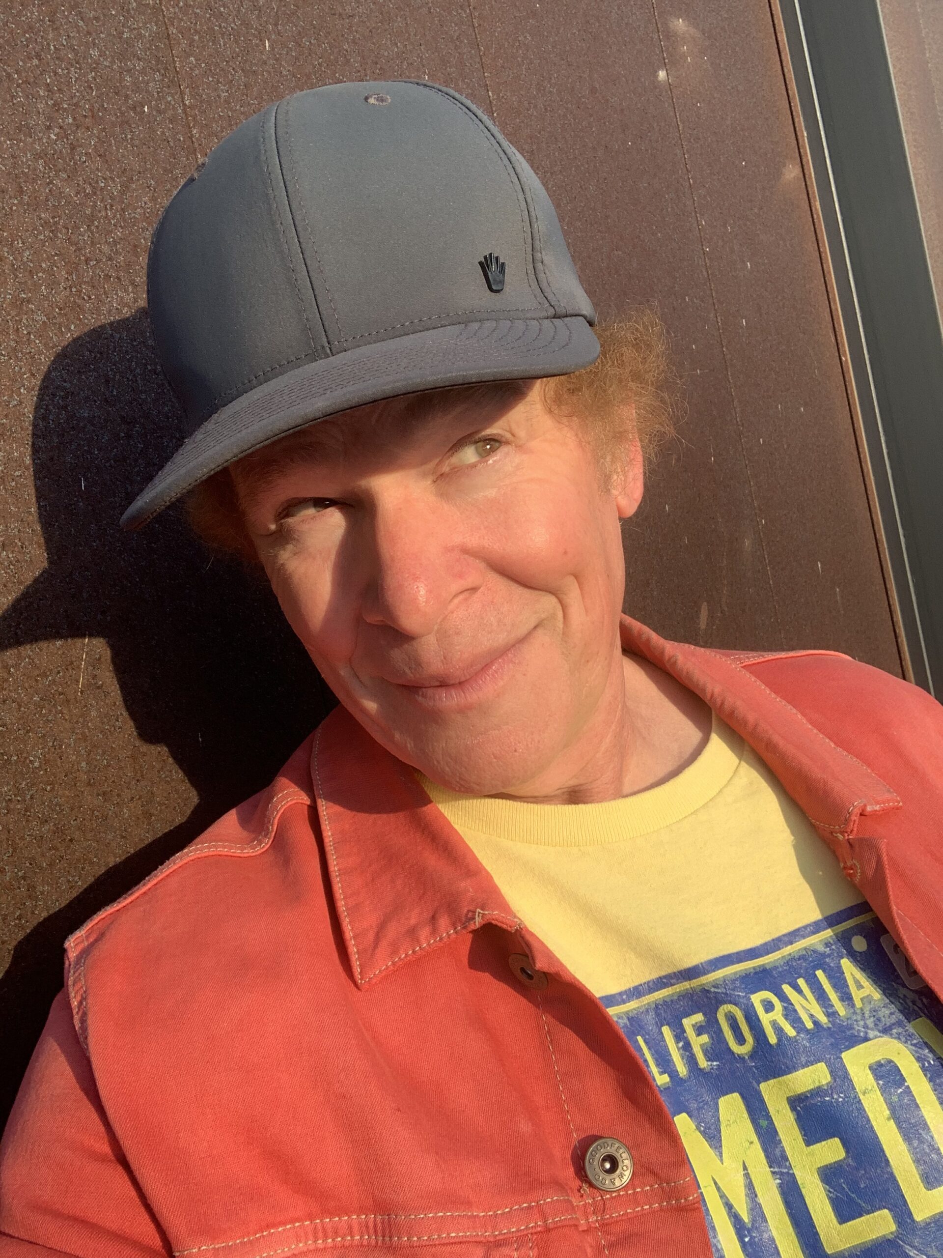 Scott headshot against a metal wall, wearing grey baseball cap, with mischievous small, looking to the upper right of photo, wearing red/orange jean jacket, and yellow tee with Comedy printed on in blue