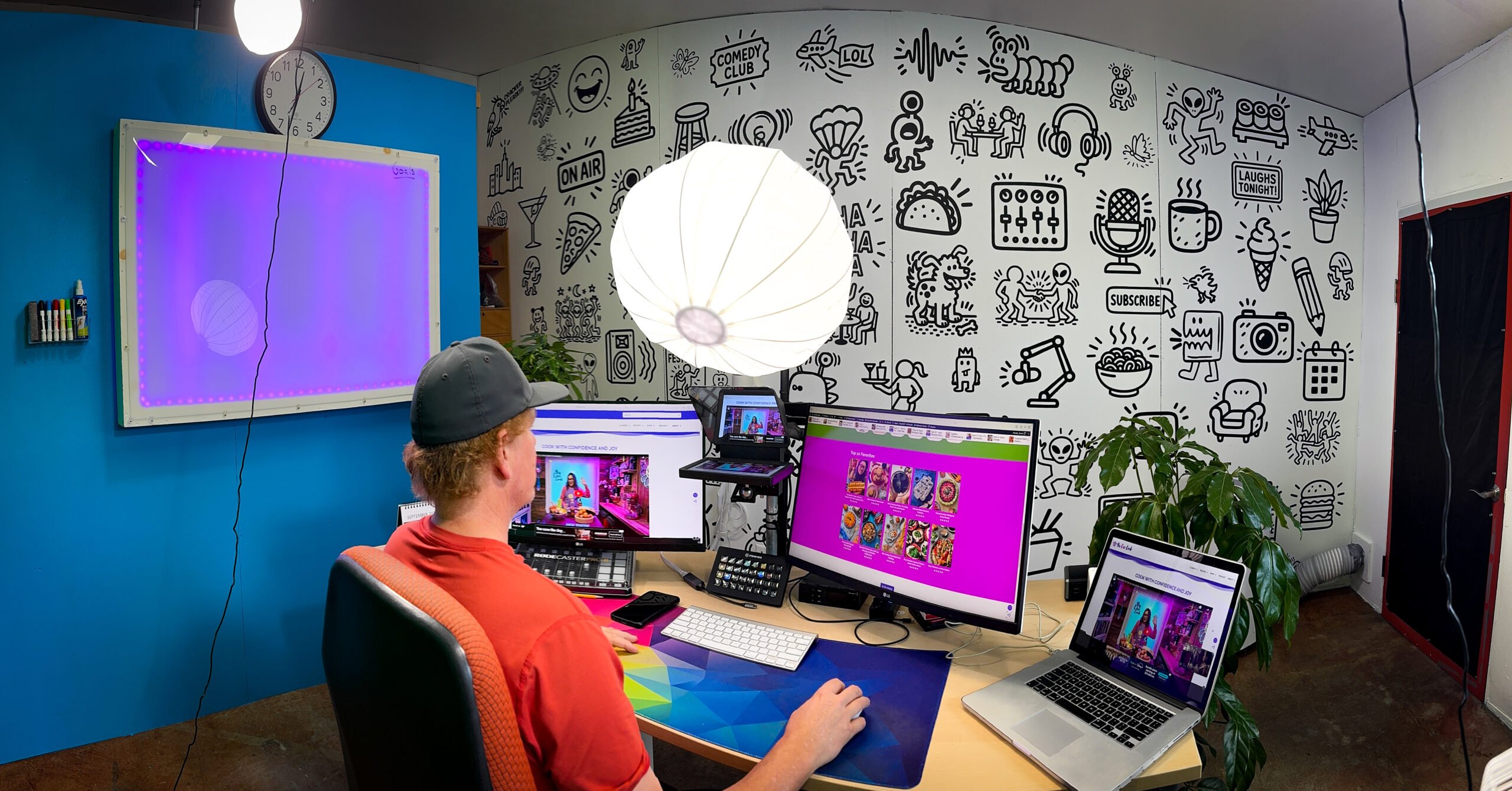 Scott sitting at table wearing orange shirt and grey baseball cap in front of his table of a laptop, and two large computer screens, and a face light, in the background is his wall covered with decals and to the left is a purple acrylic light board, and turquoise blue wall.