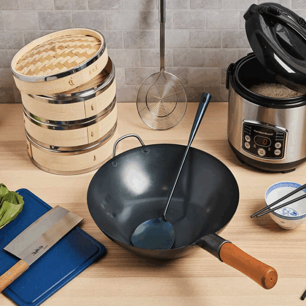 Chinese cooking equipment - steamer rice cooker, wok, spatula, cleaver, cutting board, chopsticks and rice bowl all on a butcher block counter top.