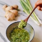 green onion ginger sauce finely chopped in oil in a rustic bowl. a wooder spoon hovers above with a spoon full of the sauce. there is a large ginger piece and 3 green onions on the marble table also