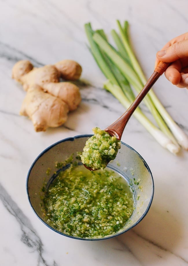 green onion ginger sauce finely chopped in oil in a rustic bowl. a wooder spoon hovers above with a spoon full of the sauce. there is a large ginger piece and 3 green onions on the marble table also